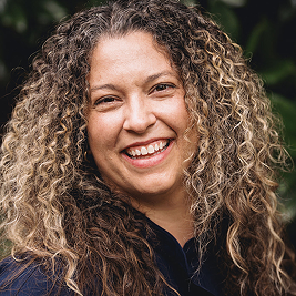 Smiling woman with long curly hair in an outdoor setting with greenery in the background.