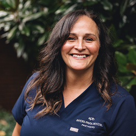 Smiling woman with long dark hair wearing a navy blue medical scrub top outdoors.
