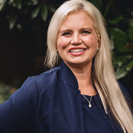 Smiling blonde woman wearing a navy blazer and necklace with greenery in the background.