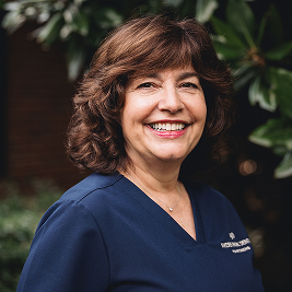 Smiling woman with curly brown hair wearing a navy blue healthcare uniform outdoors with green foliage in the background.