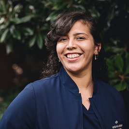 Smiling woman with curly dark hair wearing a navy blue jacket standing outdoors with green foliage background.