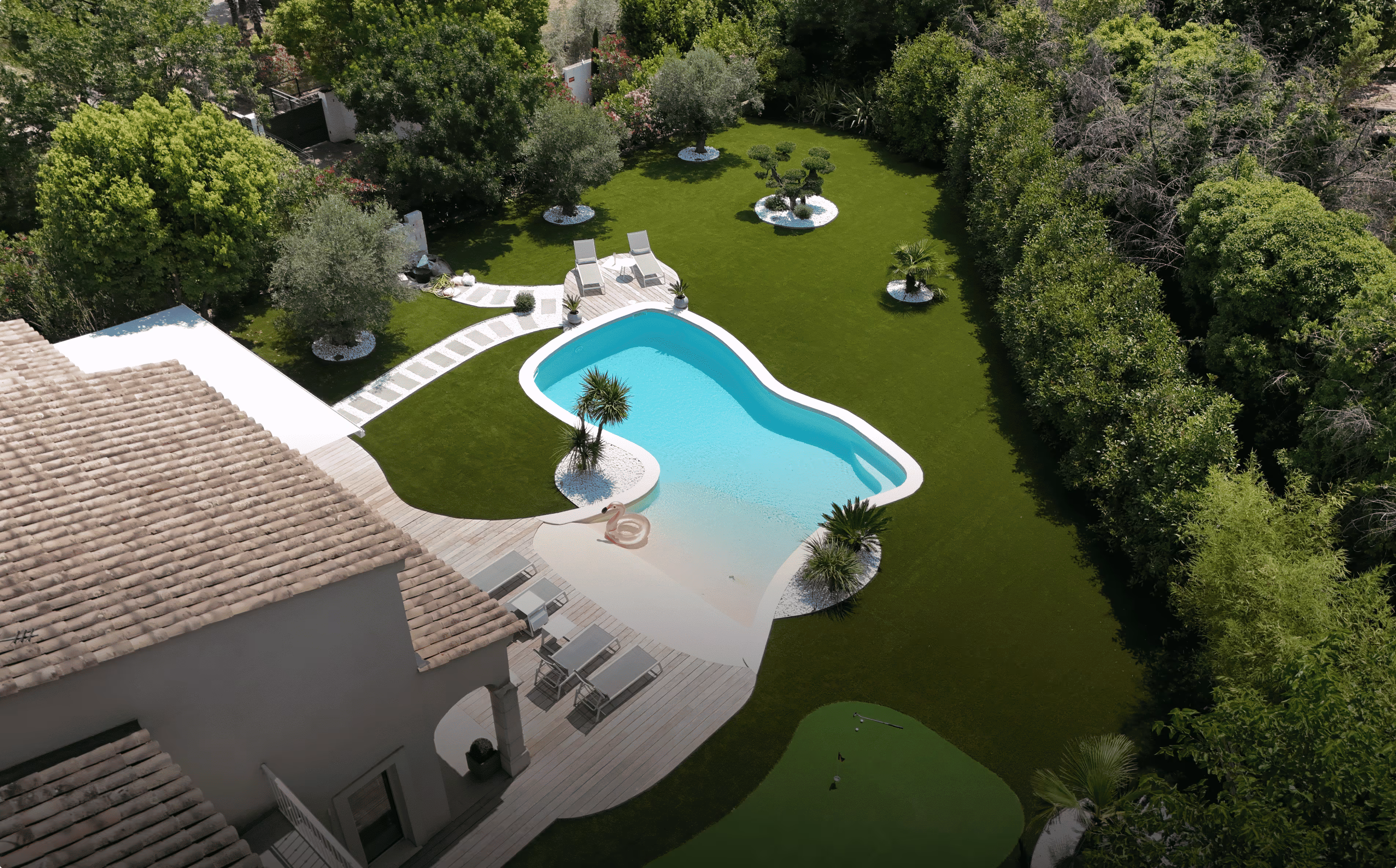 Aerial view of a backyard with a kidney-shaped swimming pool, surrounded by green lawn, trees, and lounge chairs on wooden decking.
