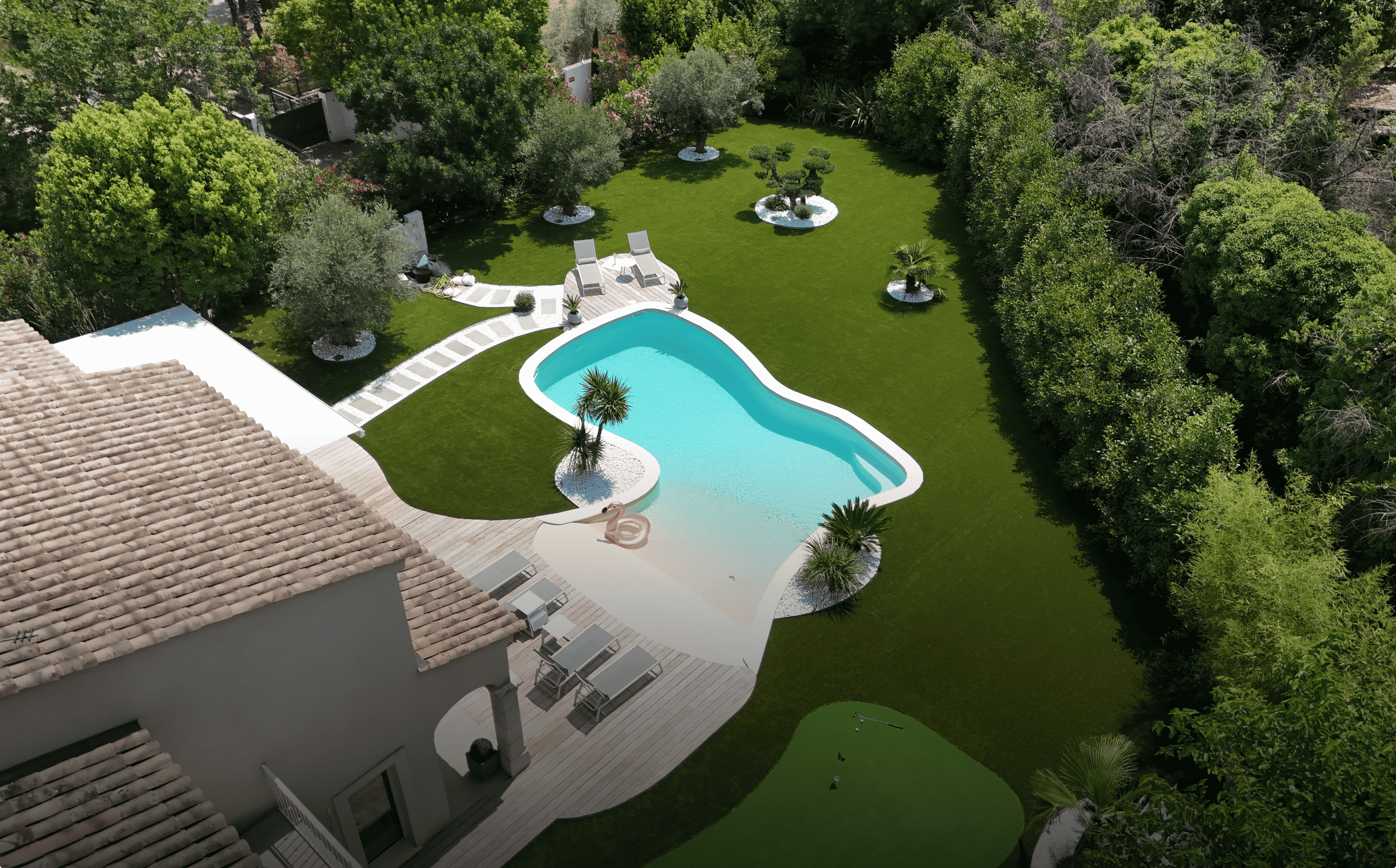 Aerial view of a backyard with a kidney-shaped swimming pool, surrounded by green lawn, trees, and lounge chairs on wooden decking.