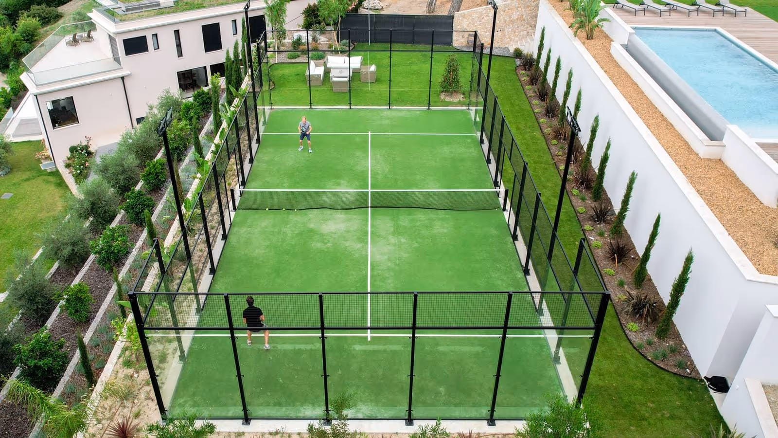 Two people playing padel on a green court with black fencing next to a modern house with a pool.