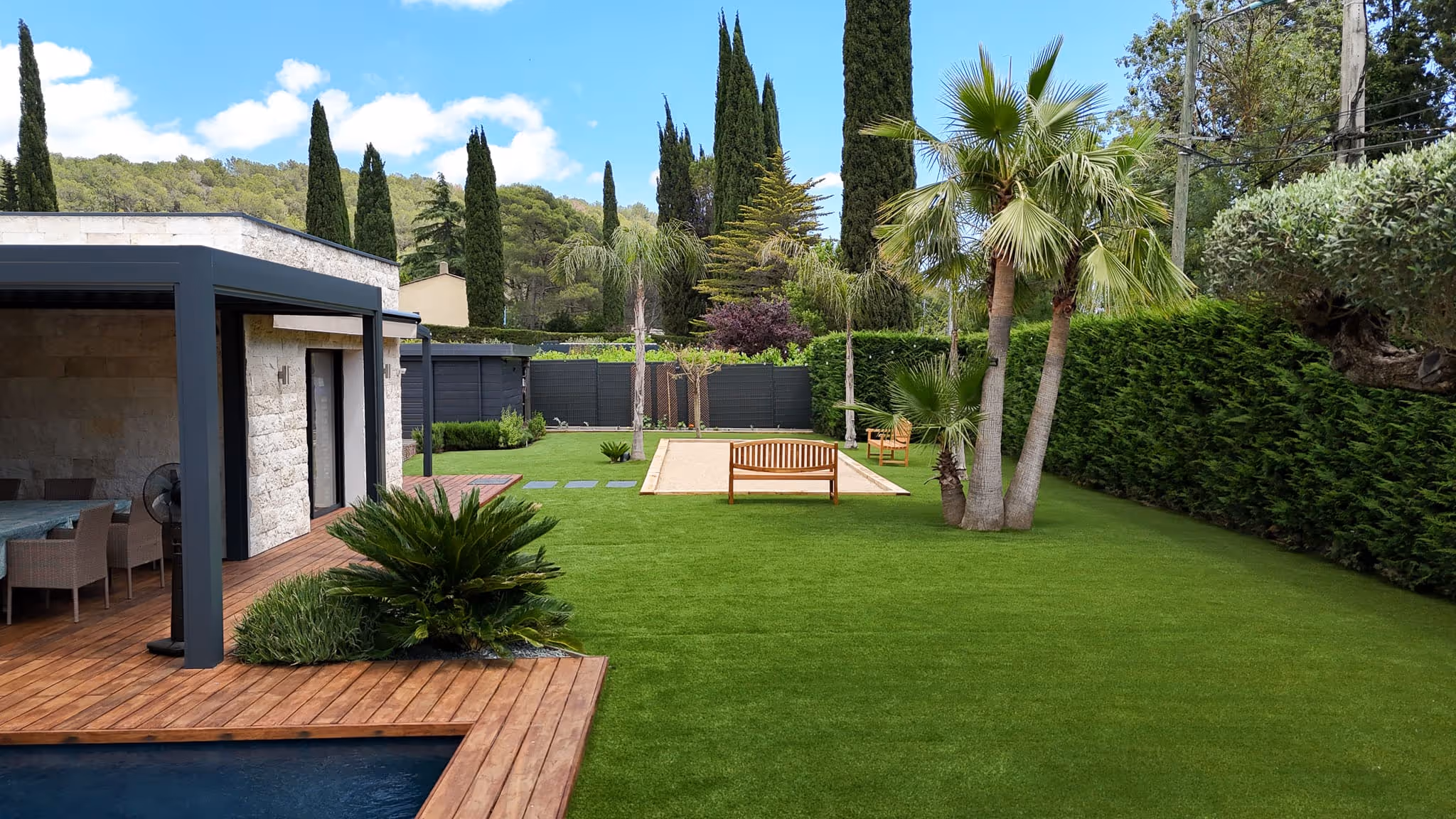 Modern backyard with a wooden deck, swimming pool, green lawn, palm trees, and benches around a sandy bocce court.