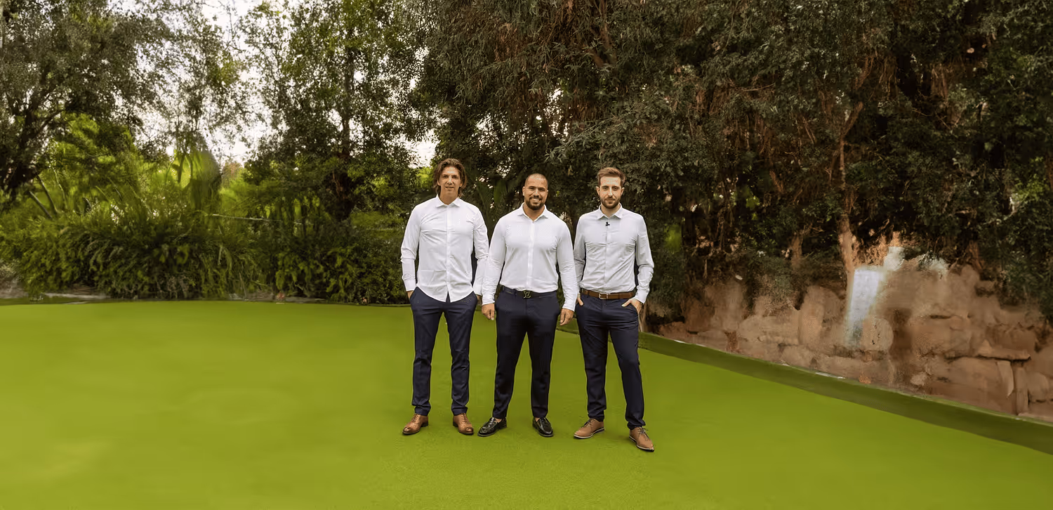 Three men standing on bright green artificial turf with trees and bushes in the background, all wearing white shirts and dark trousers.