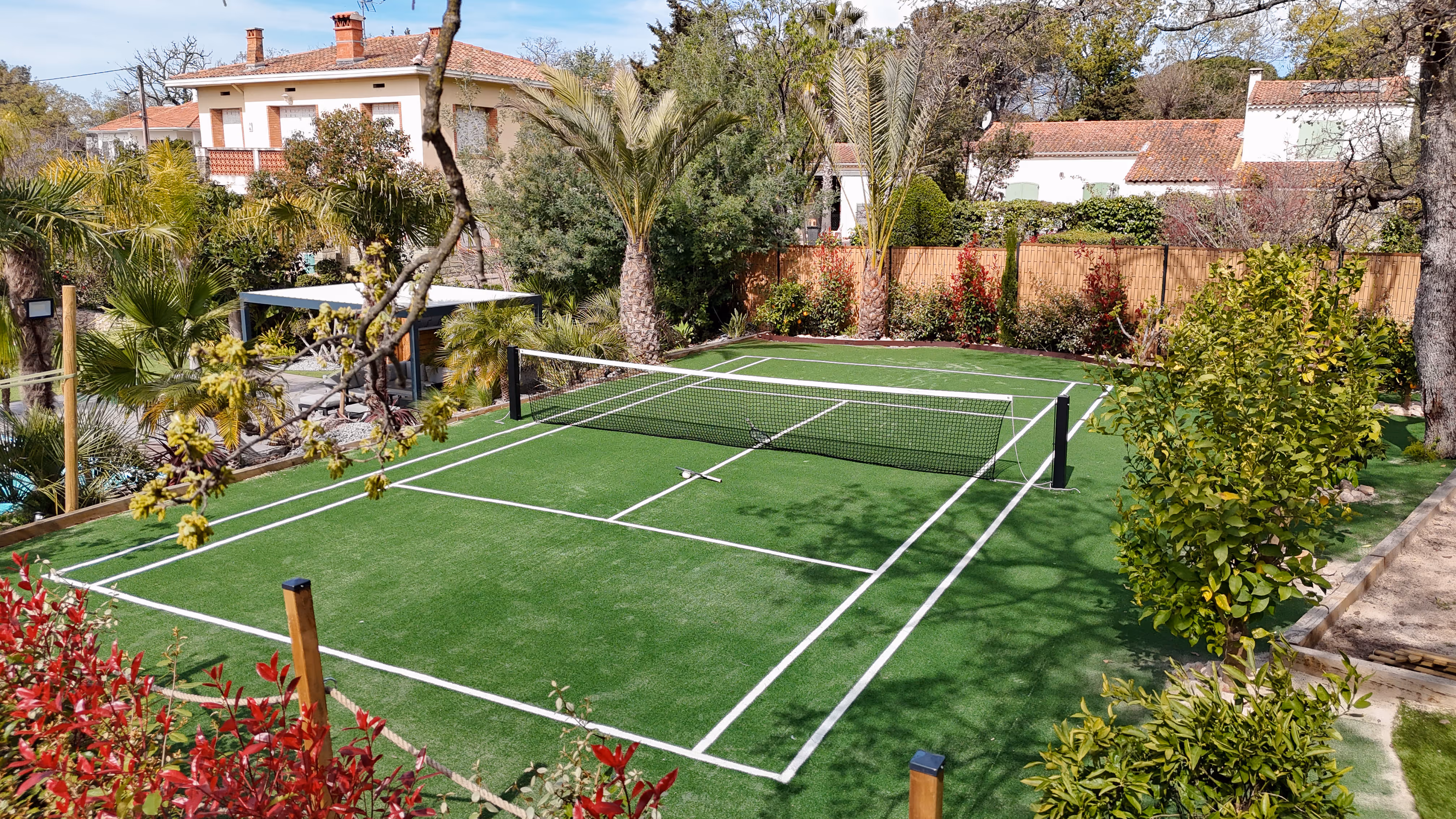 Outdoor private tennis court with artificial grass surrounded by trees and residential houses.
