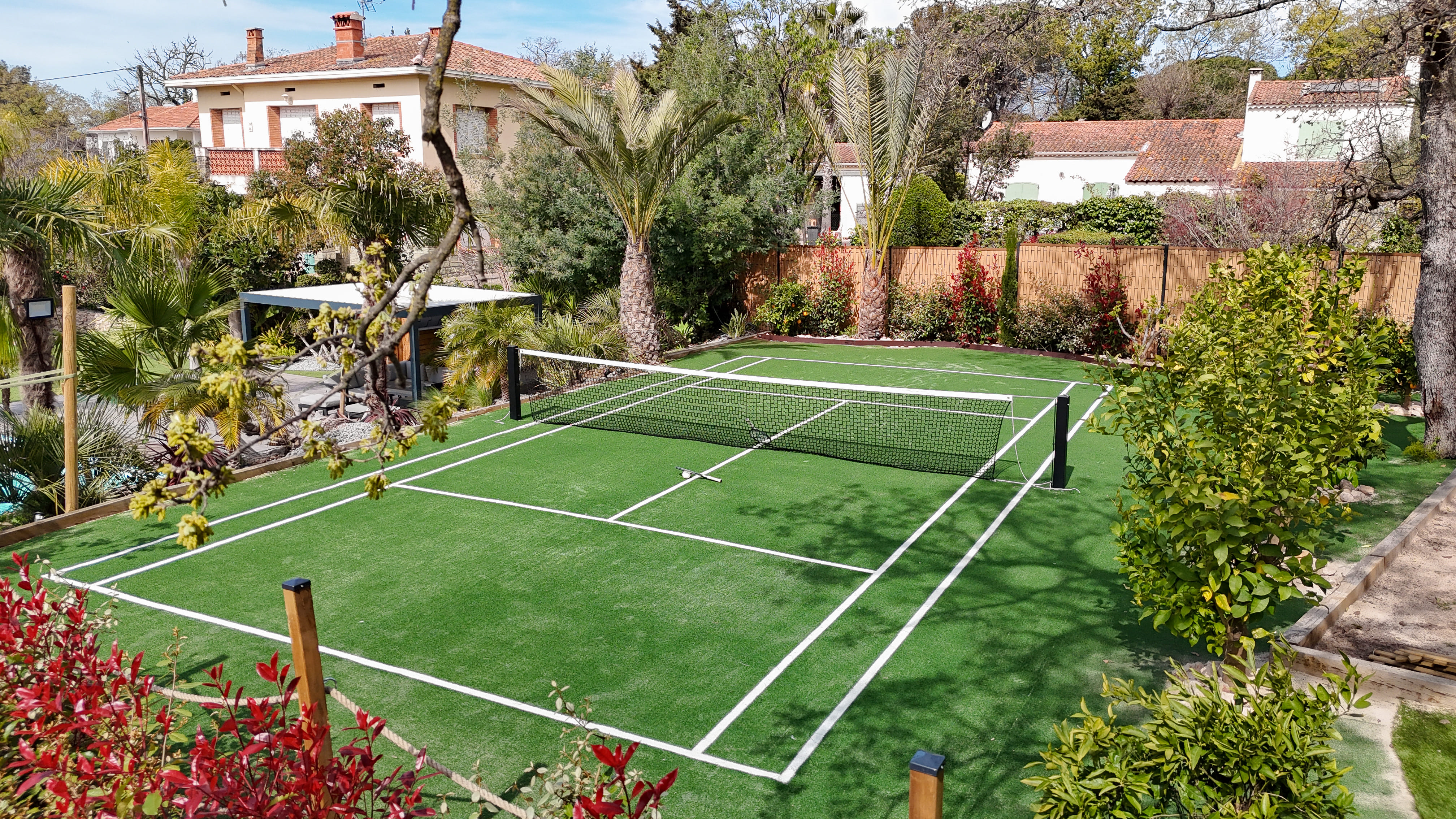 Outdoor private tennis court with artificial grass surrounded by trees and residential houses.