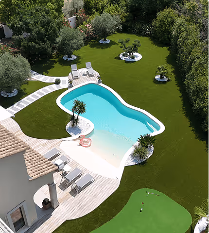 Aerial view of a kidney-shaped swimming pool surrounded by green artificial grass with lounge chairs and small palm trees, next to a house.