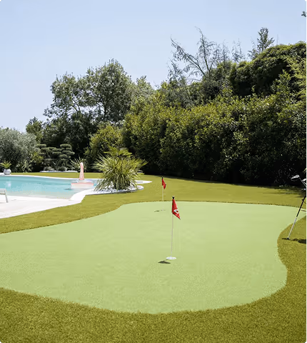 Backyard with a putting green featuring two red flags, a swimming pool, and surrounding greenery under a clear sky.