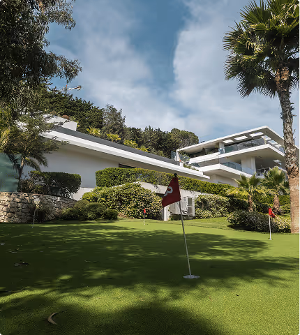 Modern house with a sloping lawn featuring golf practice flags surrounded by trees and bushes under a partly cloudy sky.