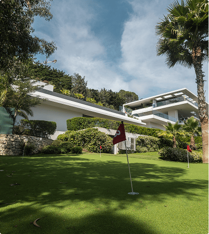 Modern house with a sloping lawn featuring golf practice flags surrounded by trees and bushes under a partly cloudy sky.