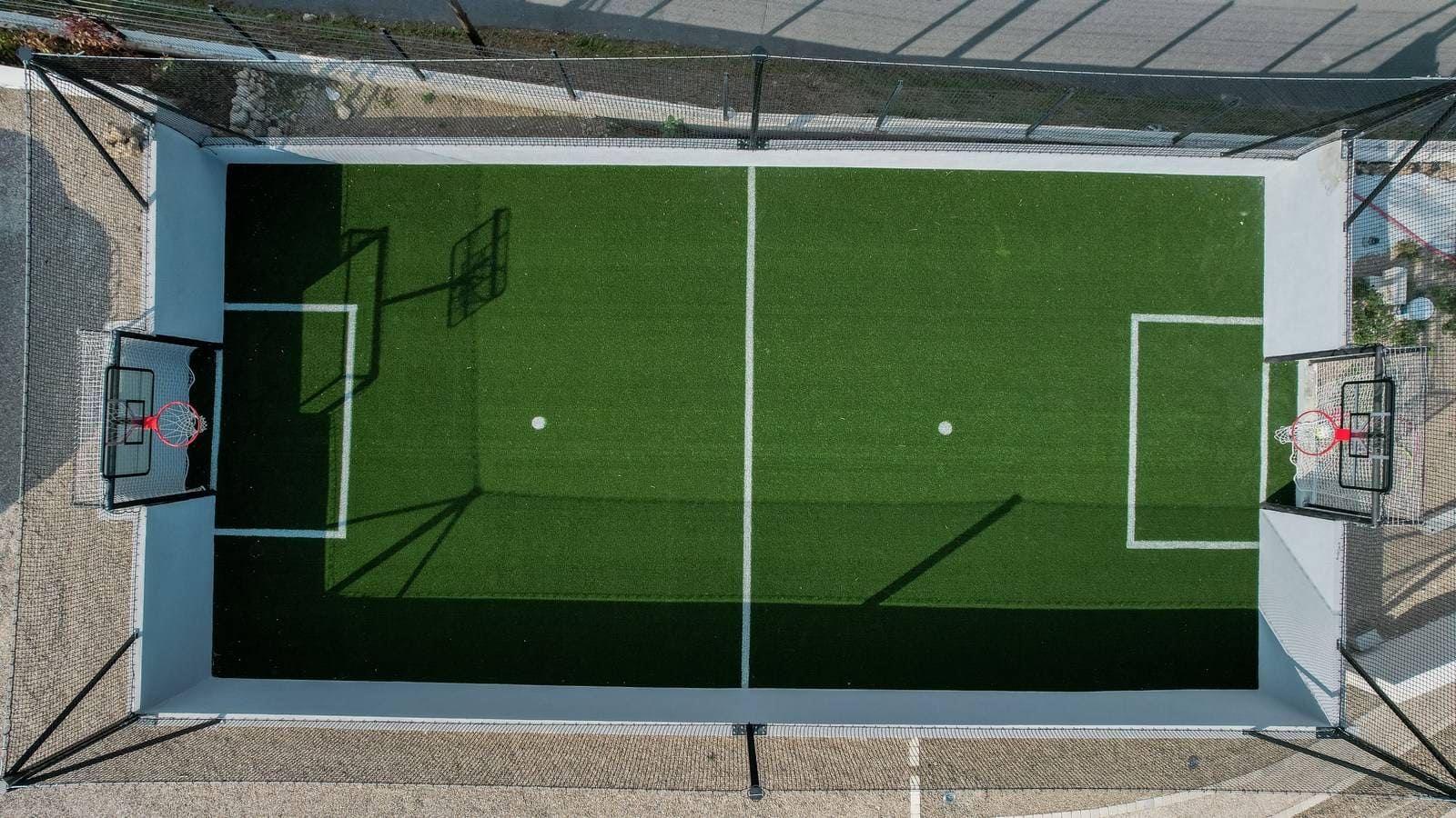 Aerial view of a small enclosed multi-sport court with two basketball hoops and a green artificial turf field marked for soccer.