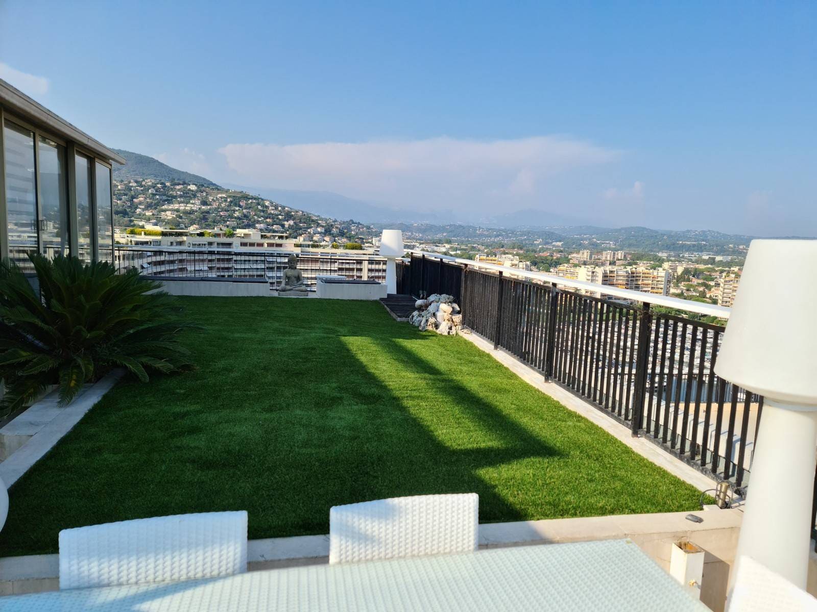Rooftop terrace with green artificial grass, white table and chairs, potted plants, and a distant view of buildings and hills under a blue sky.