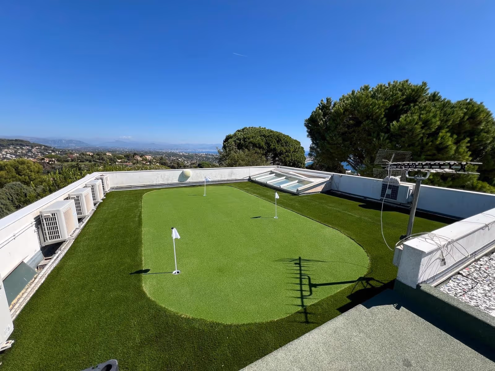 Rooftop putting green with three golf holes and flags surrounded by air conditioning units and trees under a clear blue sky.