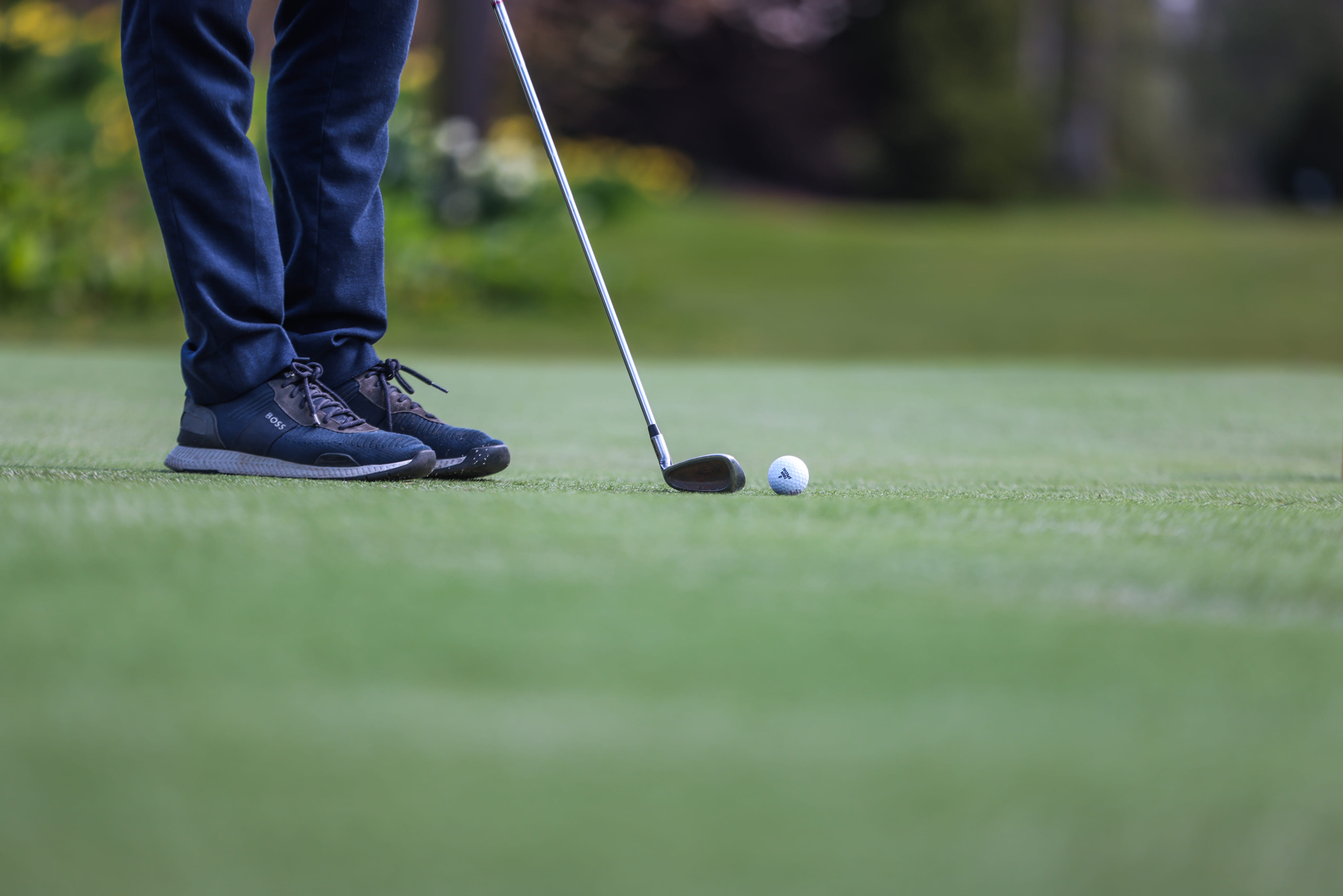 Person wearing dark blue pants and black sneakers preparing to hit a golf ball with a golf club on a green course.