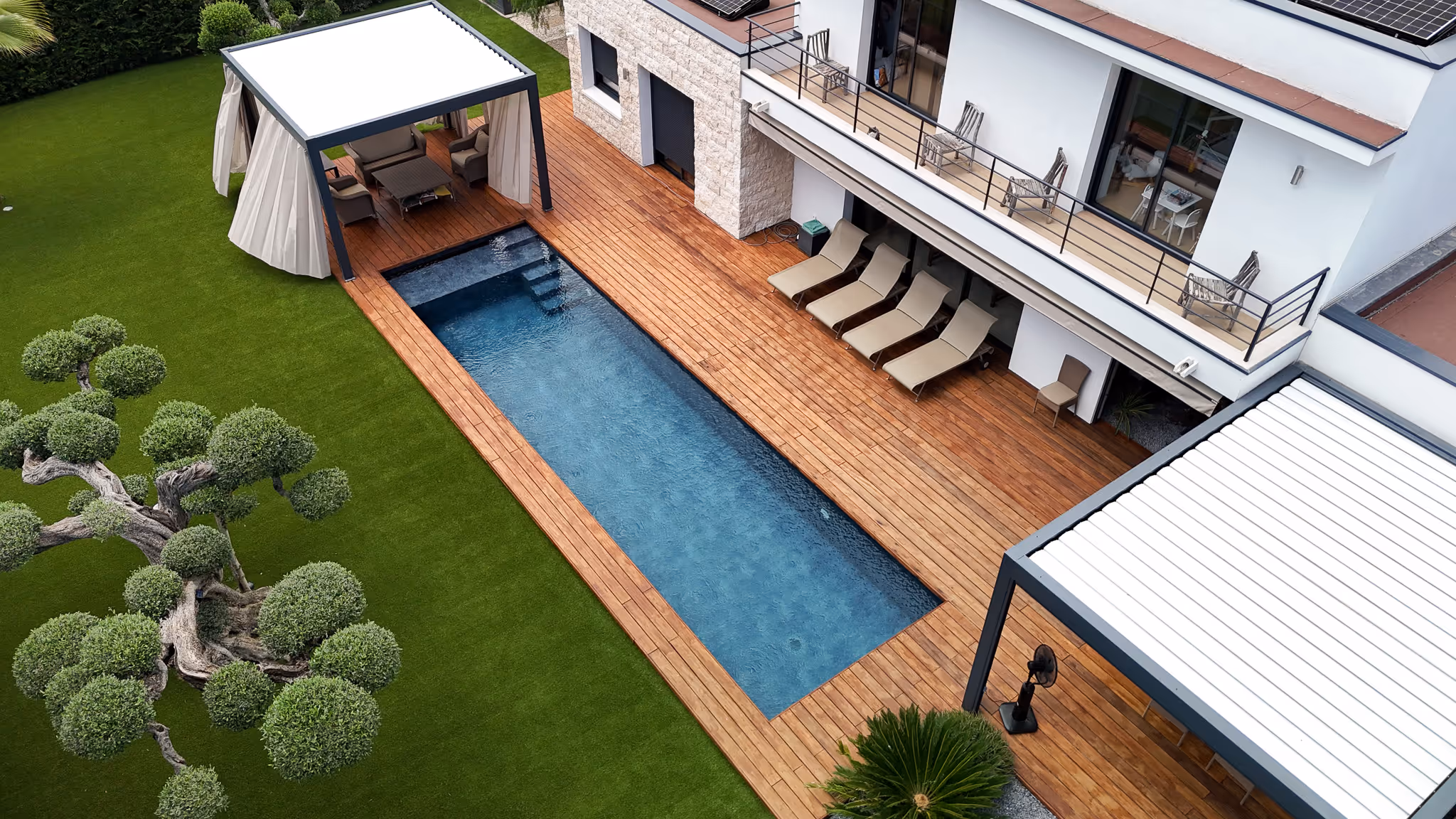 Modern backyard with a rectangular swimming pool, wooden deck, sun loungers, pergolas, and manicured green lawn with ornamental trees.