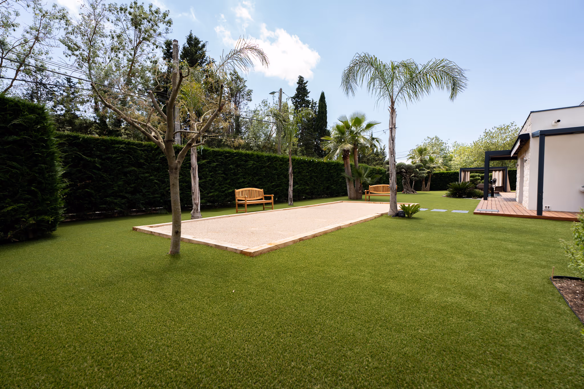 Well-maintained backyard with green artificial grass, a bocce ball court, wooden benches, palm trees, and a modern patio area attached to a house.