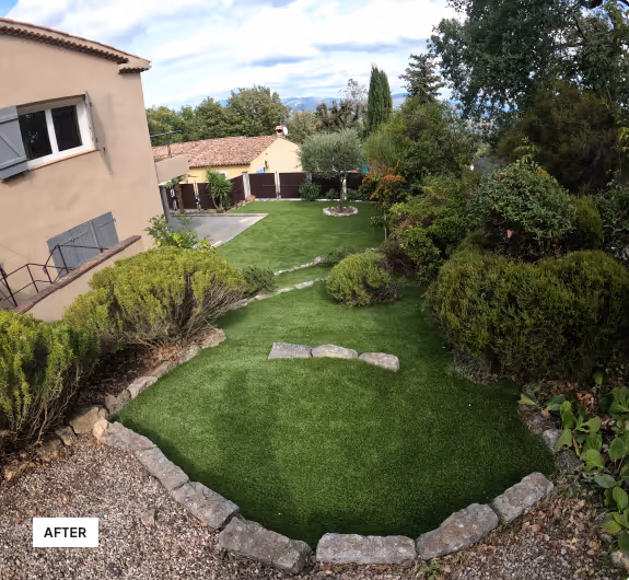 Well-maintained garden with green grass, trimmed bushes, a stone-bordered path, and a beige house with gray shutters.
