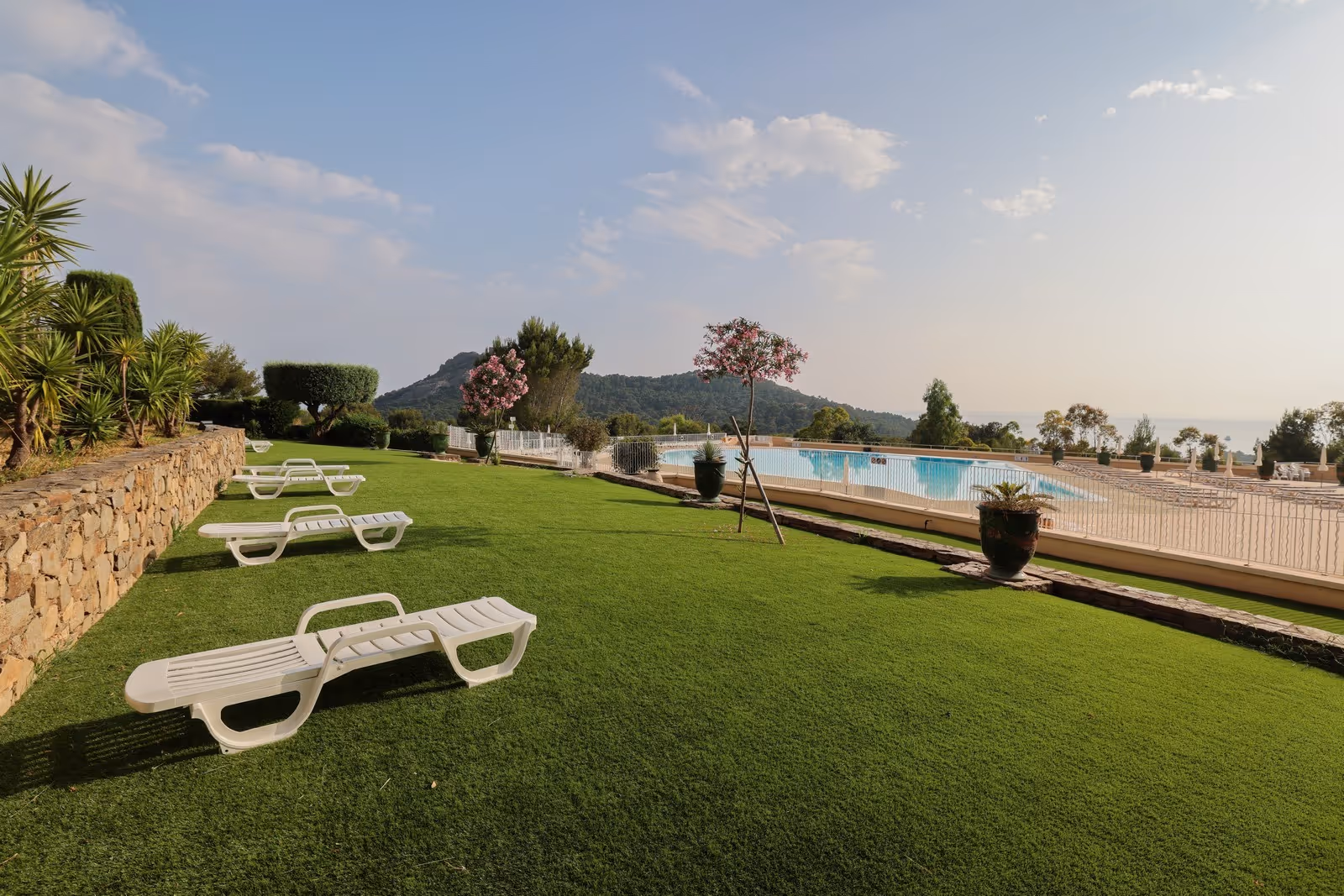 Outdoor swimming pool area with white lounge chairs on green grass and trees, overlooking hills under a partly cloudy sky.