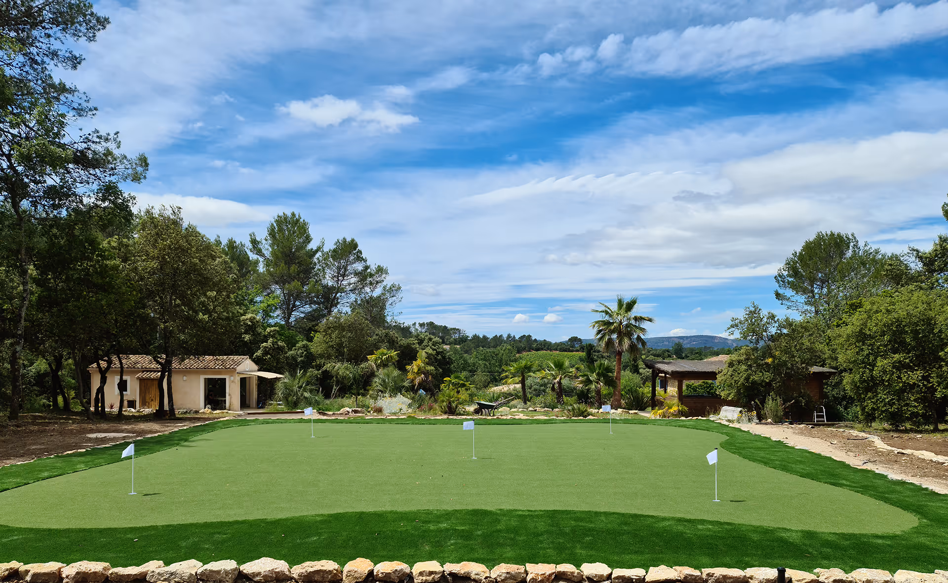 Well-maintained golf putting green with white flags surrounded by trees and small buildings under a partly cloudy blue sky.