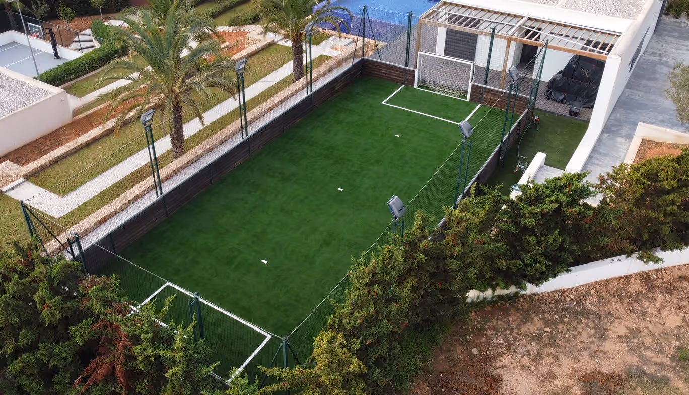 Aerial view of a small enclosed artificial grass soccer field with a goalpost, surrounded by netting, palm trees, and nearby basketball court.