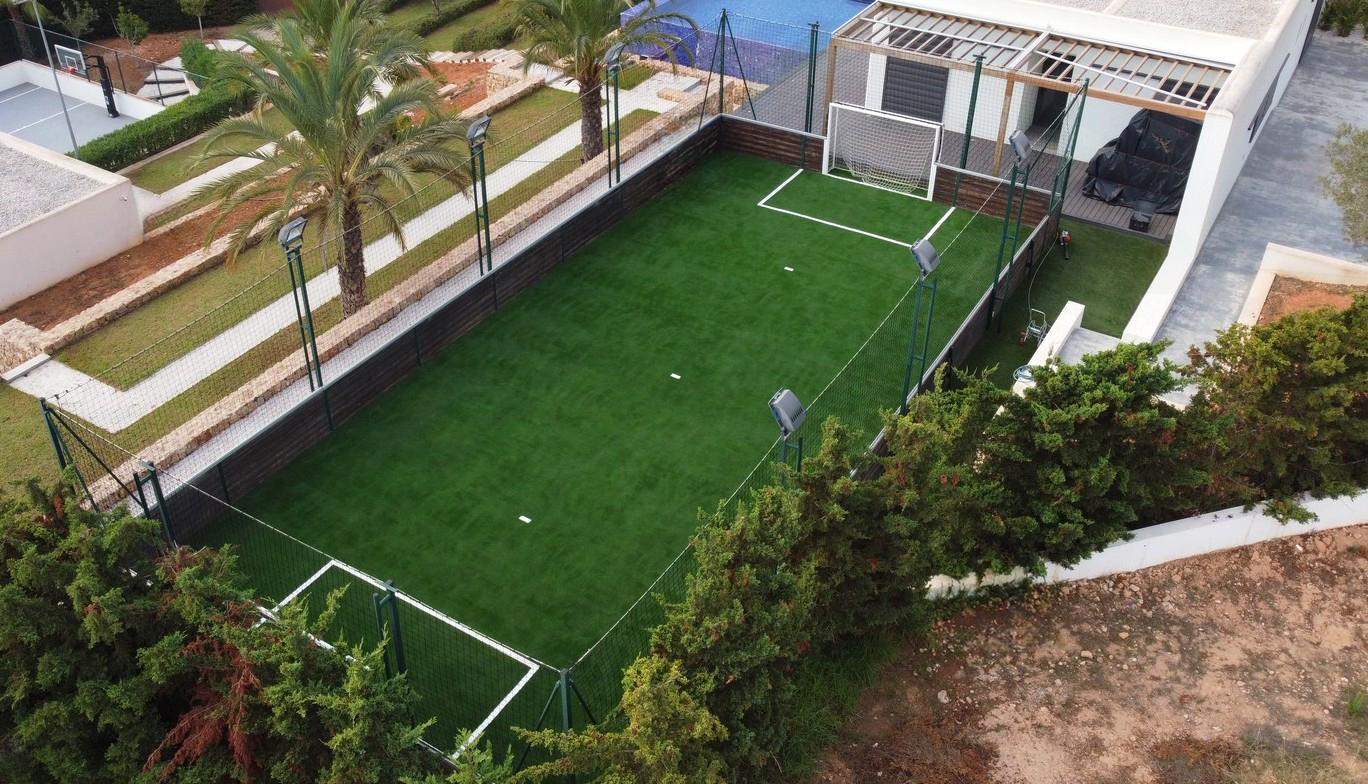 Aerial view of a small enclosed artificial grass soccer field with a goalpost, surrounded by netting, palm trees, and nearby basketball court.
