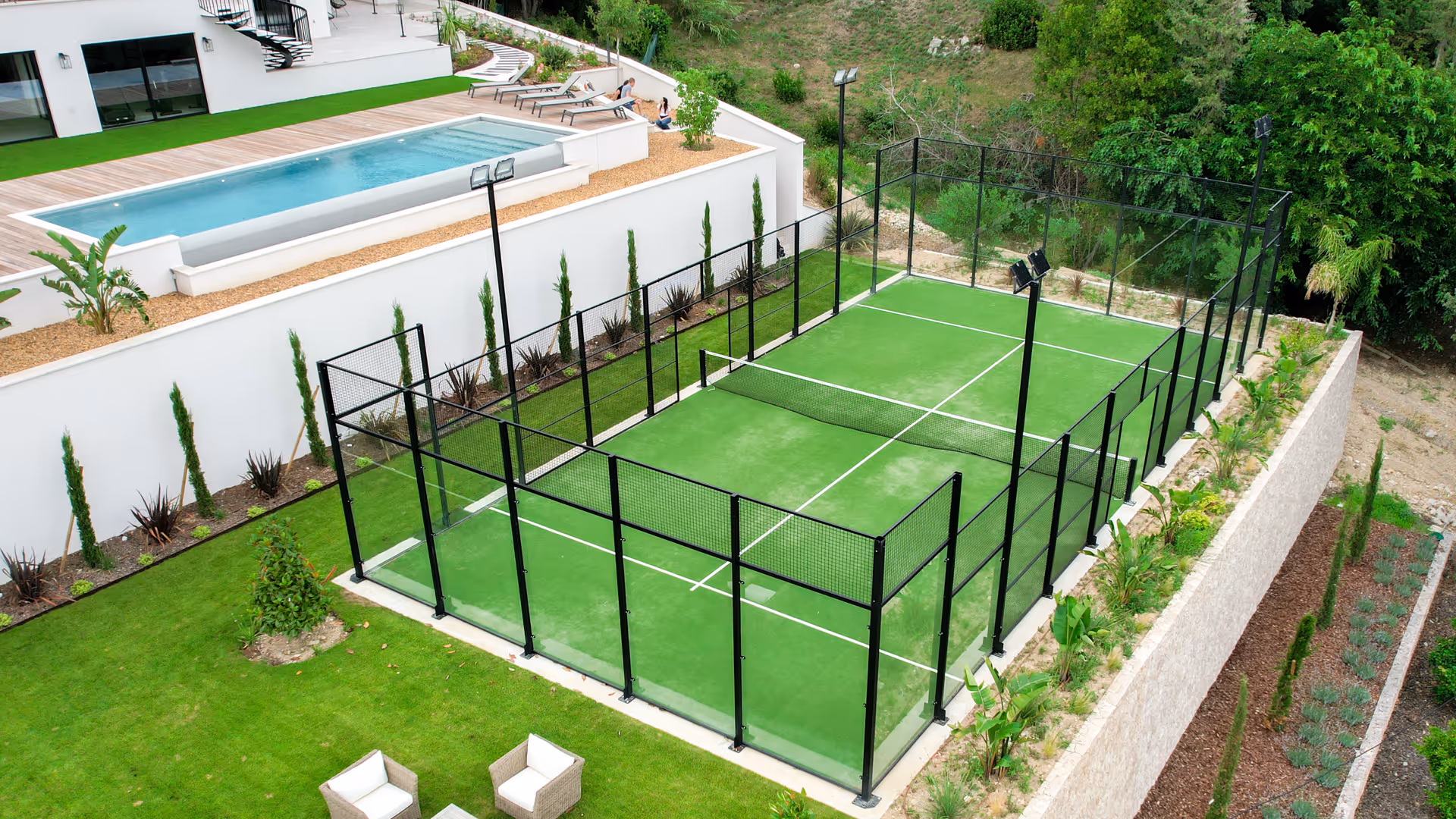 Modern outdoor paddle tennis court surrounded by glass and metal fencing next to a pool and garden area.