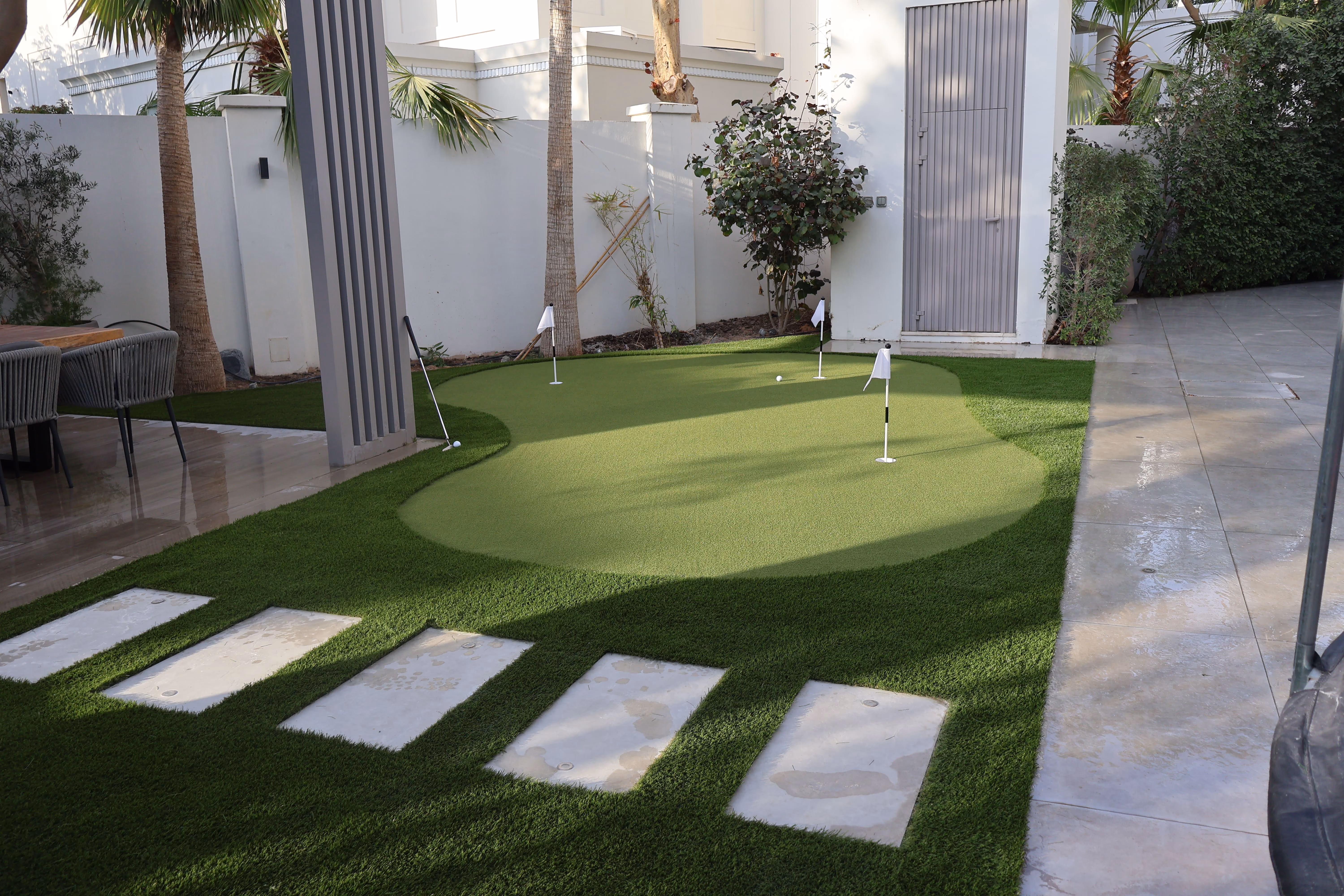 A small backyard putting green with four white flags and two golf balls on artificial grass, surrounded by a tiled patio with chairs, palm trees, and shrubs.