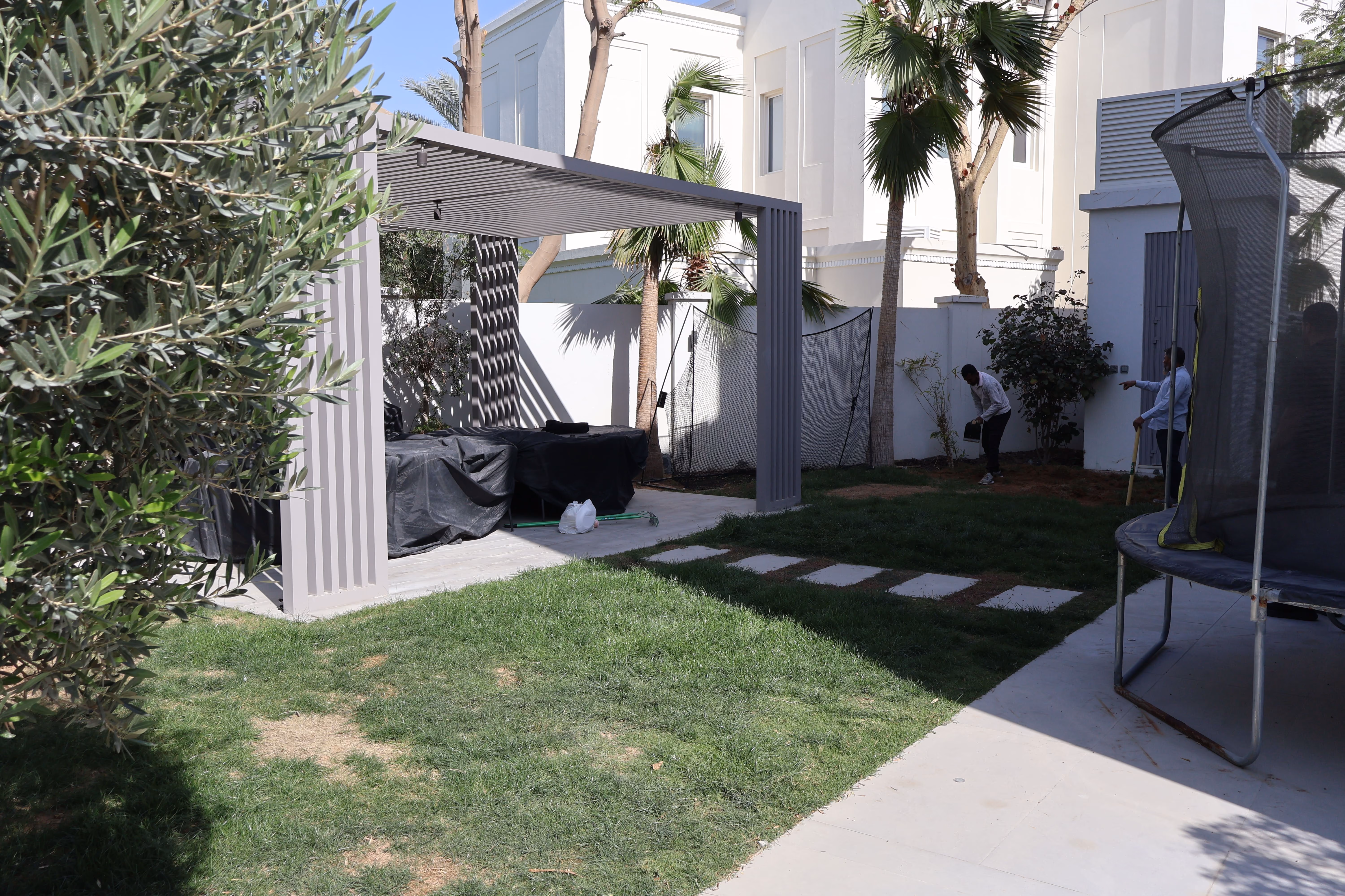 Residential backyard with green grass and patchy ground, a gray pergola covering outdoor furniture, two men working near a white wall with plants, palm trees in the background, and a trampoline on the right side on a tiled area.