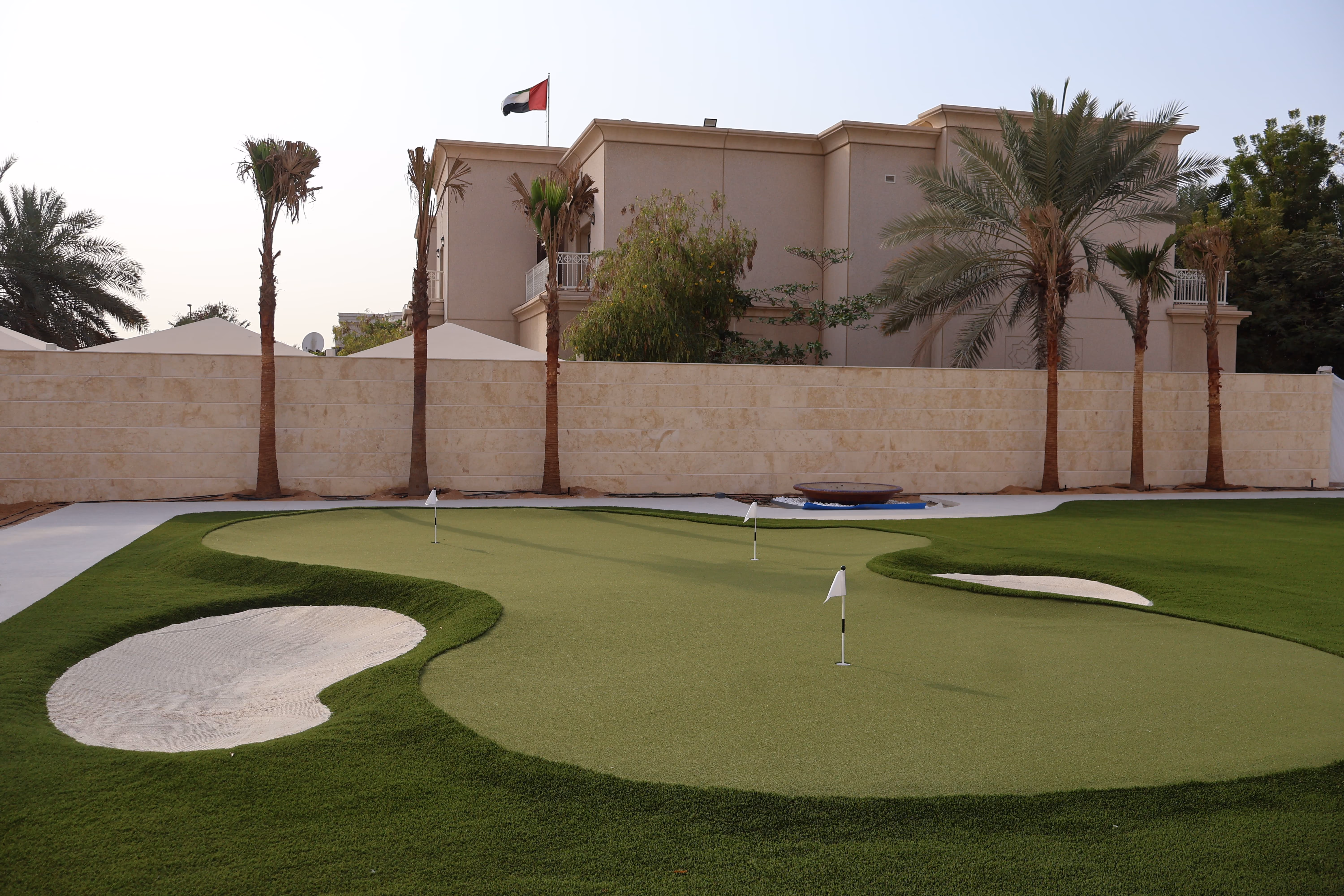 Backyard putting green with three white flags on holes, sand bunkers, and neatly trimmed grass, with a beige house, palm trees, and the UAE flag in the background.