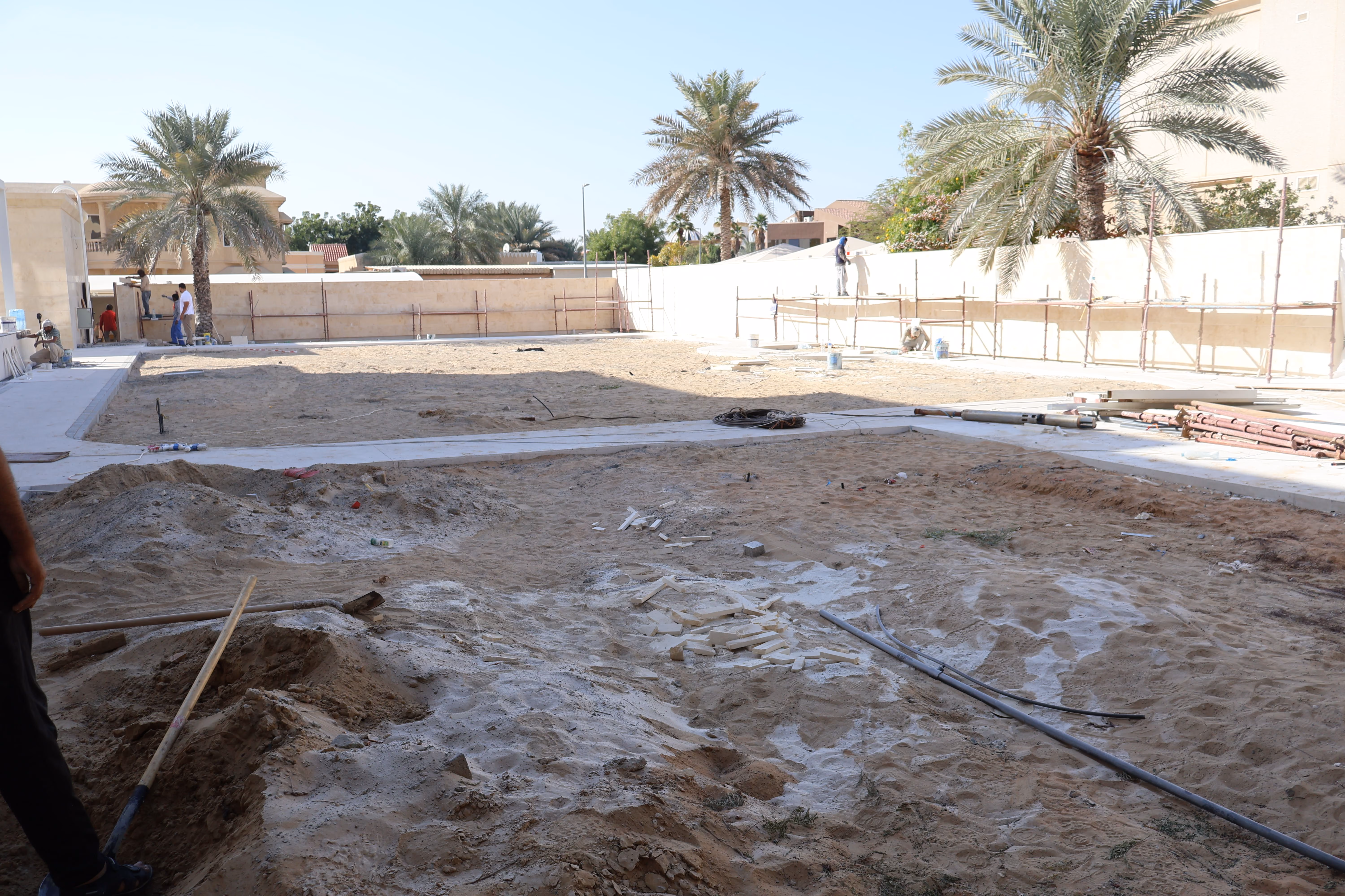 Outdoor construction site with sandy ground and several workers on scaffolding painting a beige wall surrounded by palm trees and residential buildings in the background.