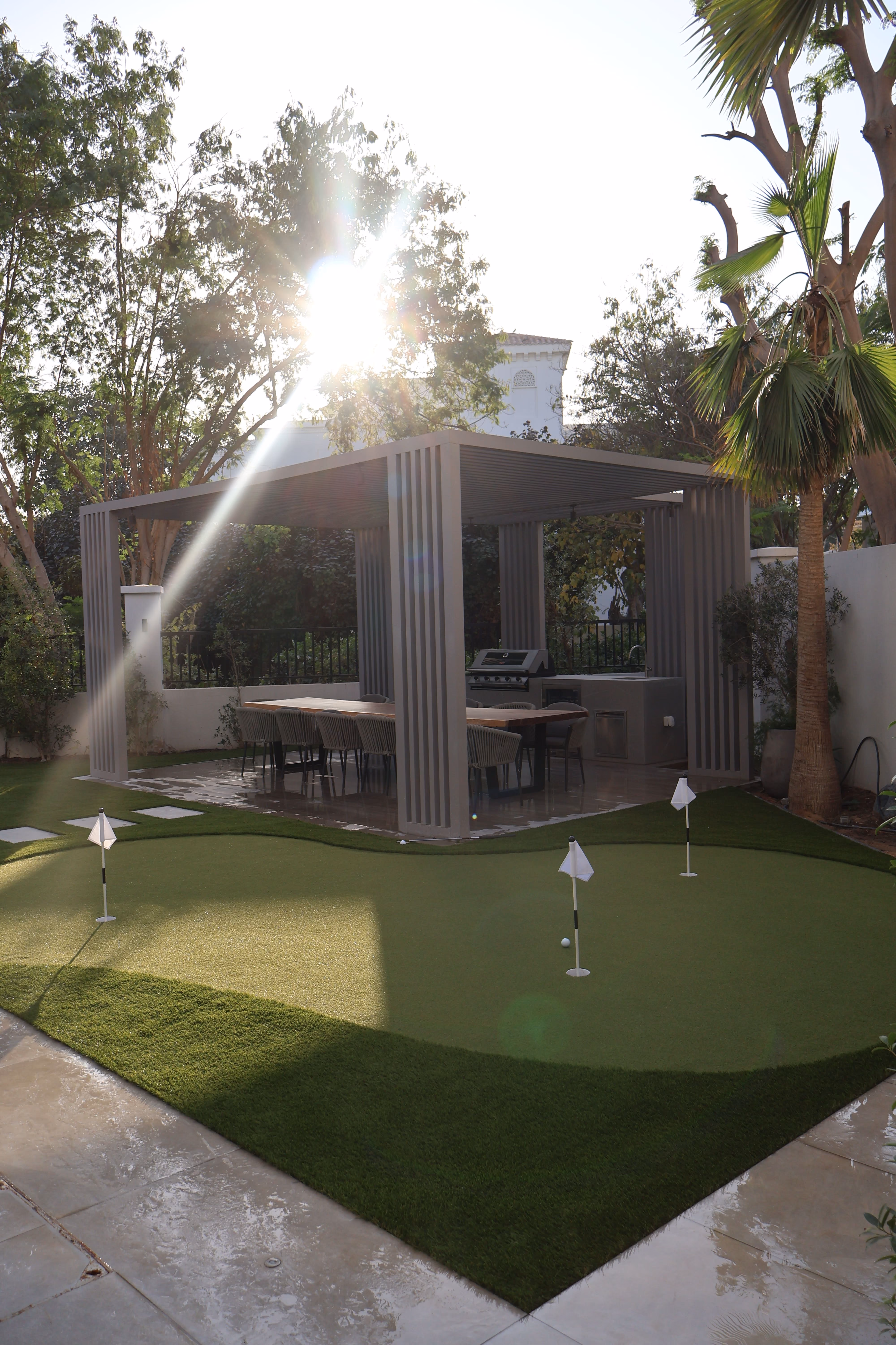 Outdoor patio with a modern pergola covering a dining table and chairs, next to a small putting green with three flags and golf balls, surrounded by trees and palm plants illuminated by sunlight.