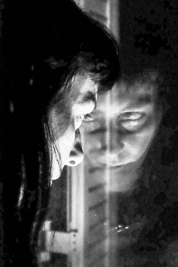 Black and white close-up of a person with long hair looking intently at their reflection in a window.