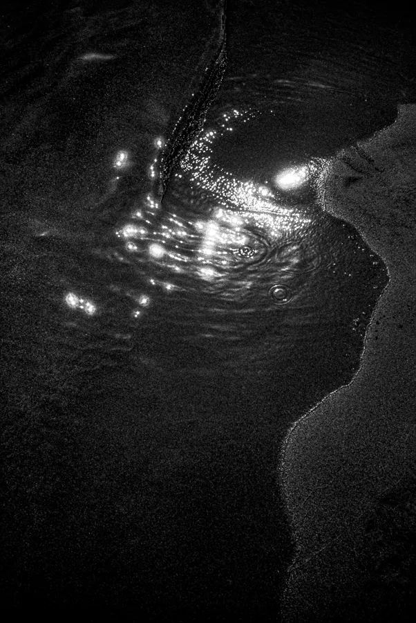 Black and white photo of water flowing between sandy banks with light reflections on the water surface.