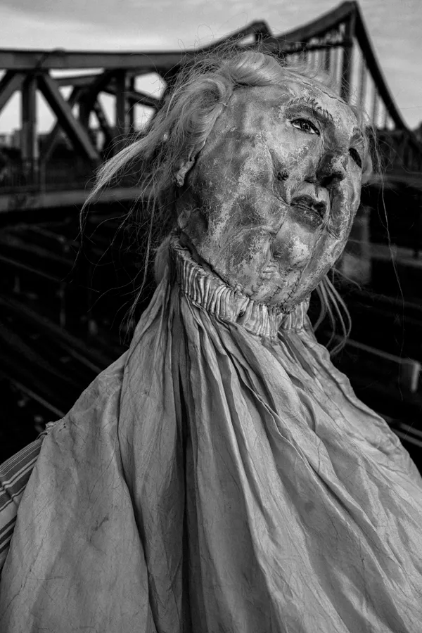 Black and white close-up of a weathered, life-sized doll or mannequin head with a wrinkled cloth body, set against a background featuring a metal truss bridge.