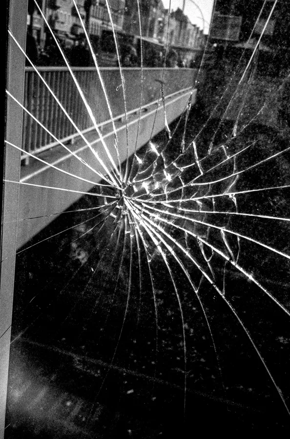 Close-up of a shattered glass window with radial cracks spreading from the impact point, showing a blurred street and railing in the background.