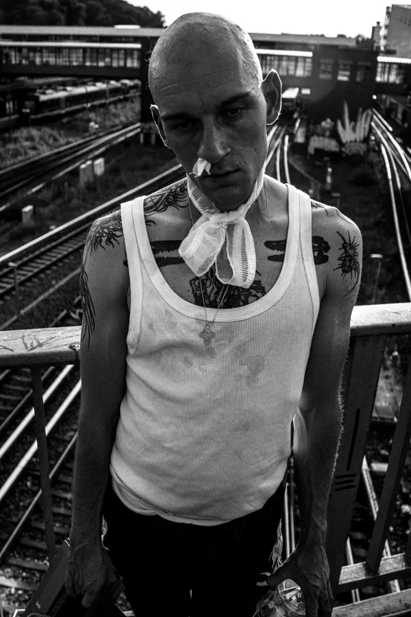 Black and white portrait of a bald man with tattoos wearing a white tank top and a gauze tied around his neck, standing on a bridge over train tracks.