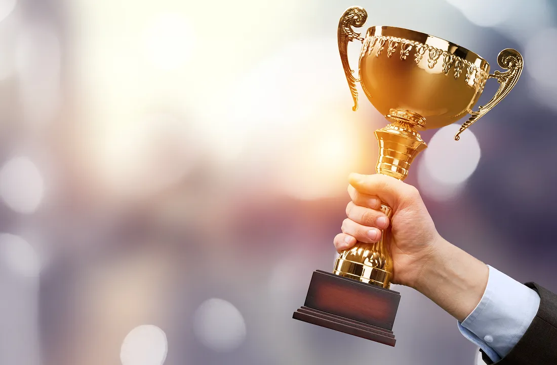 Hand holding a golden trophy cup with ornate handles against a blurred background.