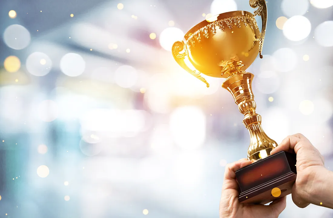 Hands holding a golden trophy cup against a blurred background with light bokeh effects.