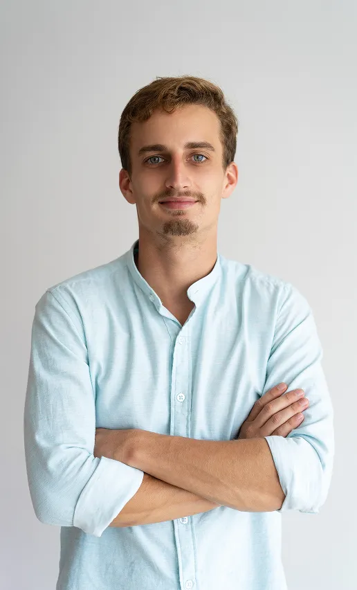 Young man with light brown hair and blue eyes wearing a light blue shirt with sleeves rolled up, standing with arms crossed and smiling slightly.
