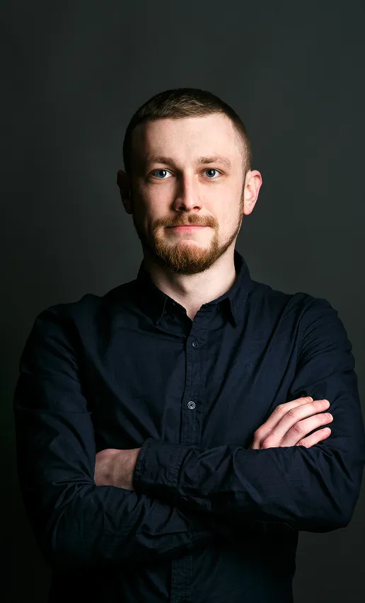 Young man with short hair and beard wearing a black button-up shirt, standing with arms crossed against a dark background.