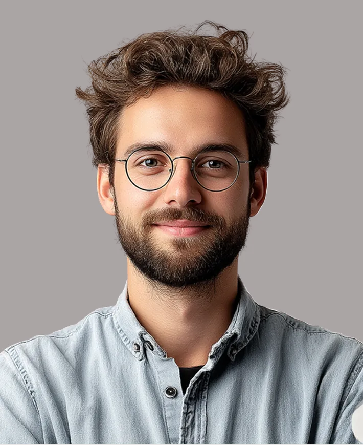 Smiling young man with curly hair, beard, round glasses, and light denim shirt against a plain background.