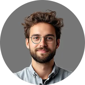 Smiling young man with curly hair, beard, and glasses wearing a light blue shirt against a gray background.