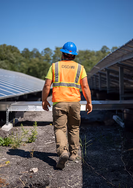 Engineer wearing DSDJ safety vest and blue helmet walking through solar panel installation site.