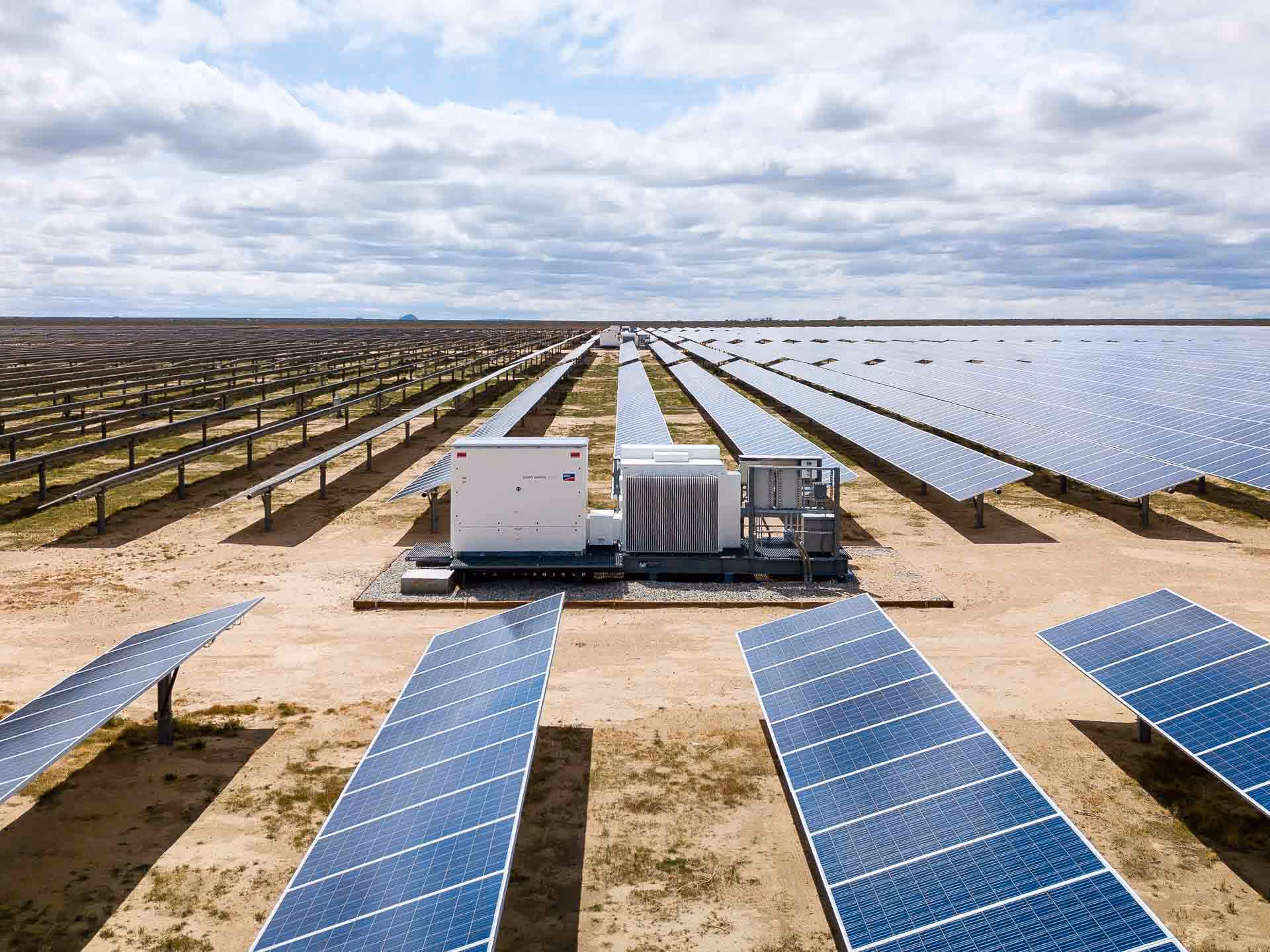 Large utility-scale solar farm with rows of photovoltaic panels and central inverter equipment under a cloudy sky.
