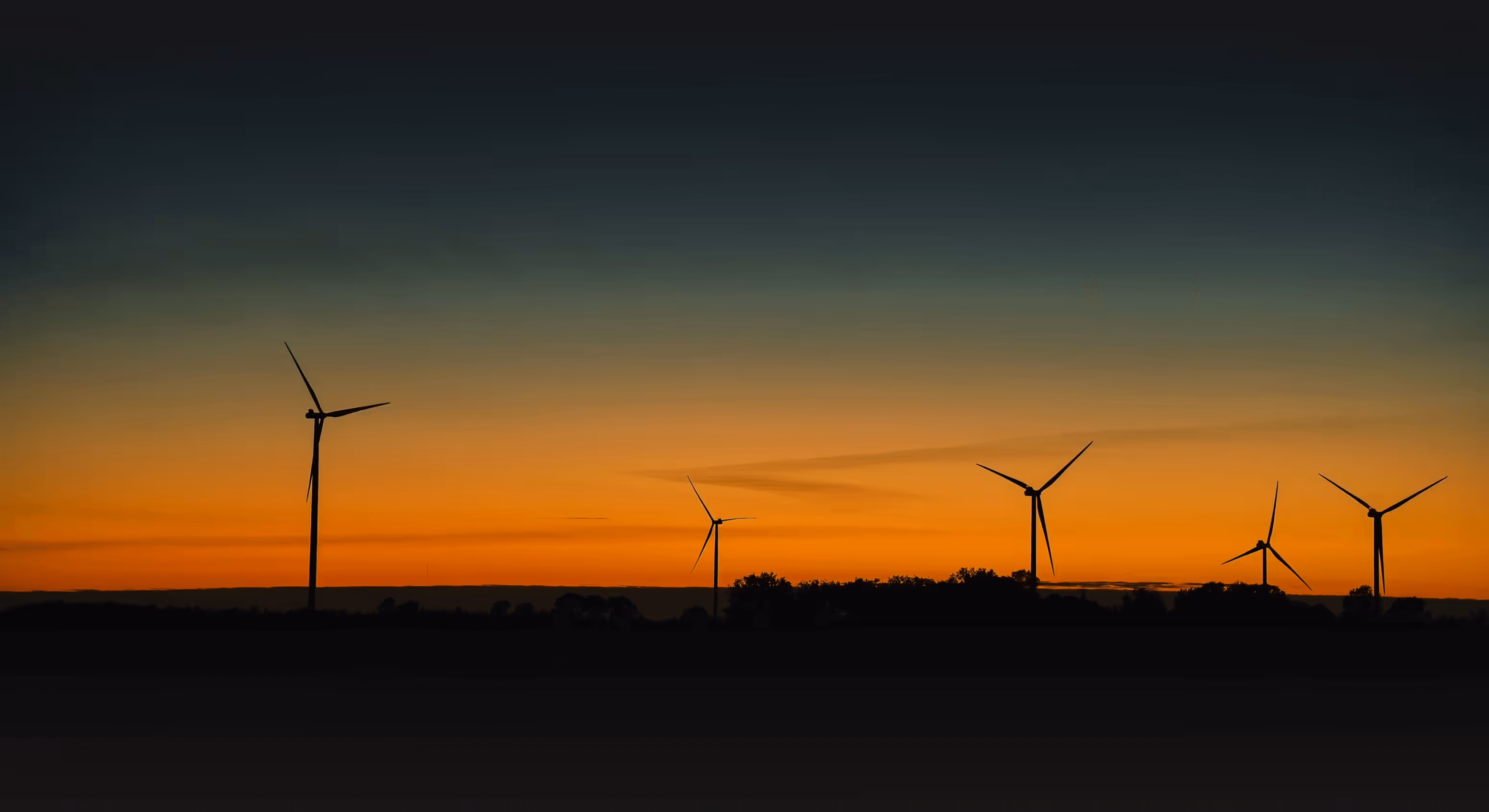 Silhouettes of five wind turbines against a gradient sunset sky with shades of orange and blue.
