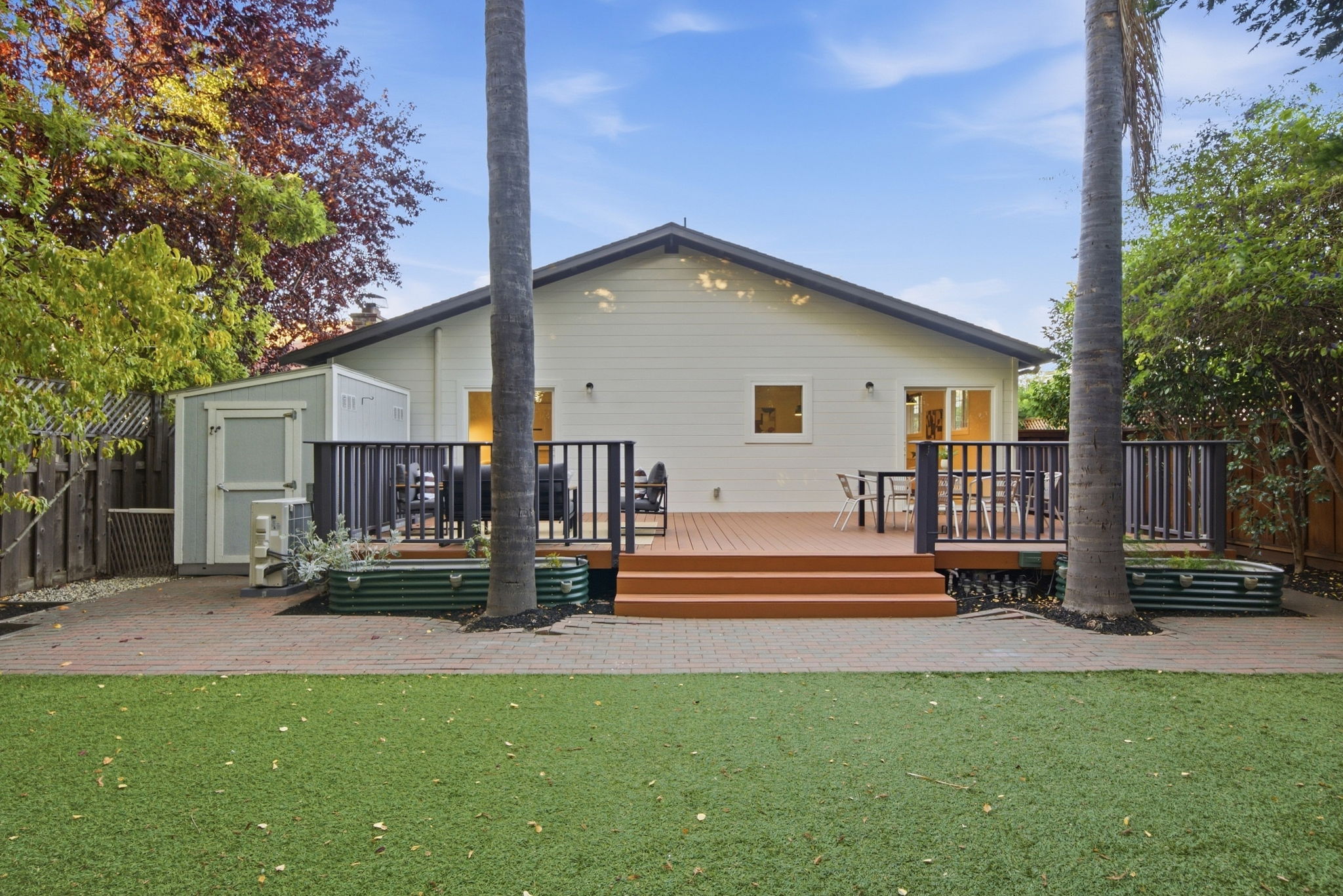 Backyard of a single-story house with a brown wooden deck featuring two large palm trees, outdoor seating areas, and a small light gray storage shed to the left, surrounded by green artificial grass and trees with colorful foliage under a clear blue sky.