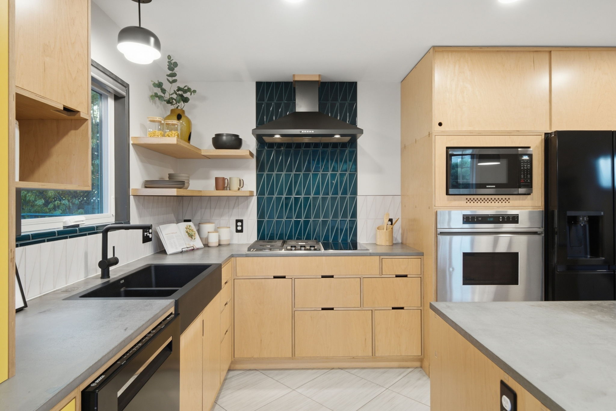 Modern kitchen with light wood cabinets, gray concrete countertops, a black sink and faucet below a window, open shelves with dishes and decor, a stainless steel oven and microwave, a black refrigerator, and a blue geometric tile backsplash behind a black range hood and gas cooktop.