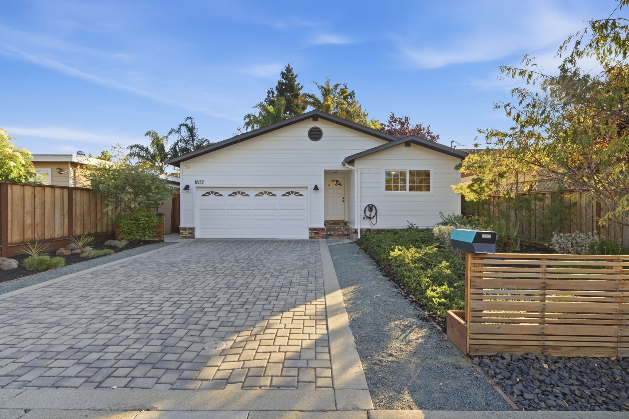 Single-story white house with a three-car garage, paved driveway, and a small garden area with a modern wooden fence and mailbox in front under clear blue sky.